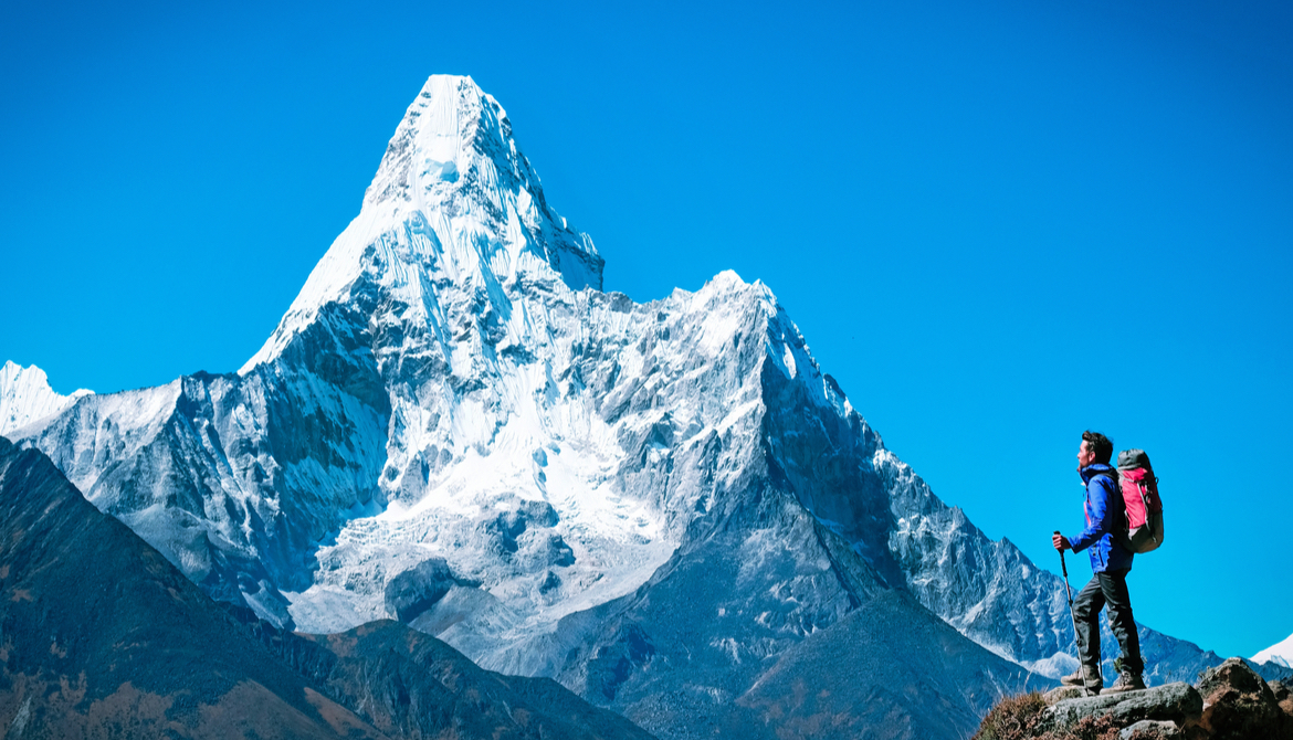 Hiker at summit of mountain in the foreground looking toward an even higher mountain peak in the background