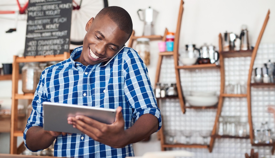 Café small business owner smiling and speaking on phone while using digital tablet