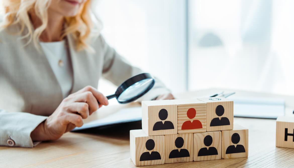 Female HR professional examines pyramid of wooden blocks representing job candidates with a magnifying glass