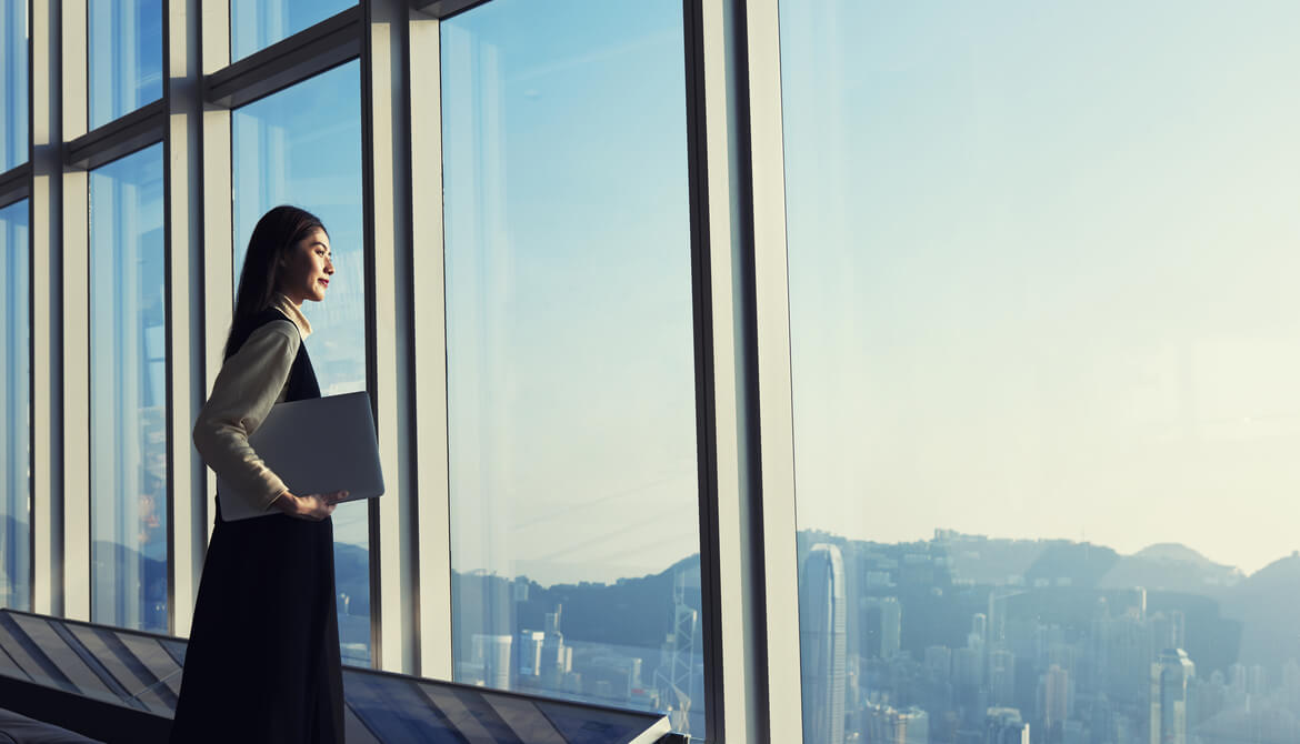 Young Asian millennial businesswoman holding laptop looks out high office windows