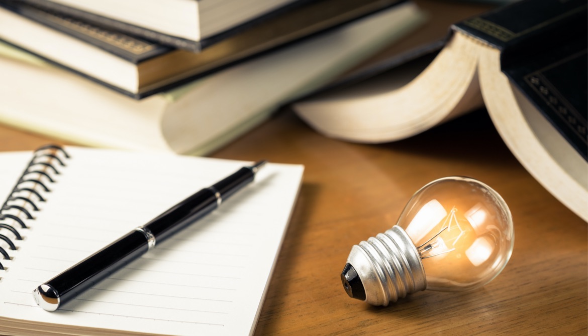 Small light bulb glowing on the desk with notebook and many books on background