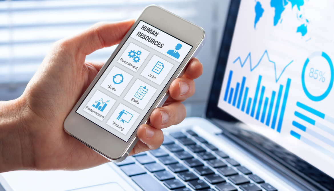 HR professional holds smartphone with human resources app while looking at reports on laptop