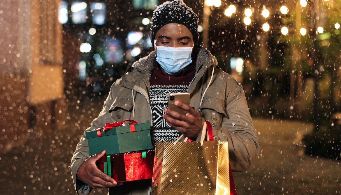 African American man wearing mask looks at smartphone while carrying holiday shopping in snow