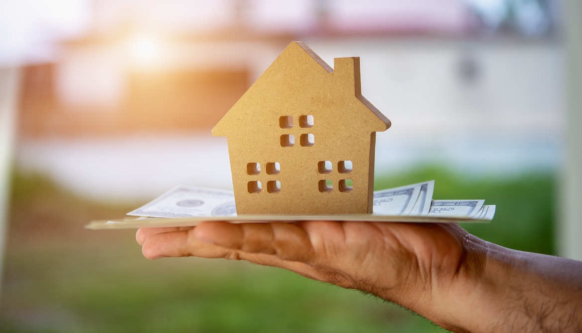 wooden cut-out of a house sitting on a stack of US dollars on an outstretched hand