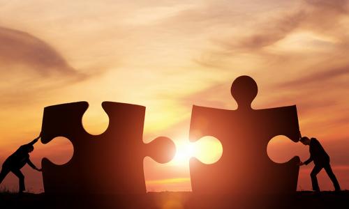 two business men push together two jigsaw puzzle pieces against a backdrop of a sunset