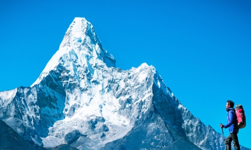 Hiker at summit of mountain in the foreground looking toward an even higher mountain peak in the background