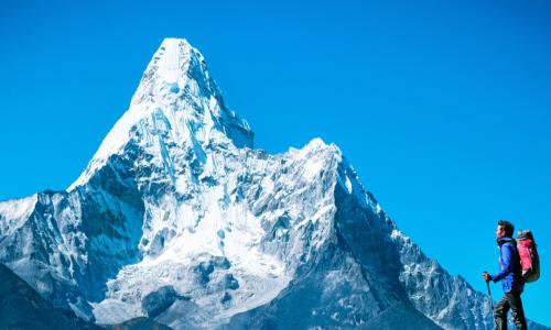 Hiker at summit of mountain in the foreground looking toward an even higher mountain peak in the background