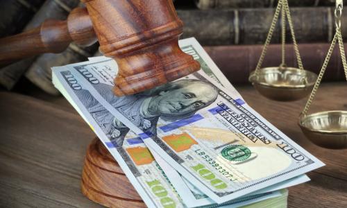 Wooden gavel resting on a stack of $100 bills in front of a set of brass scales representing compensation and regulation