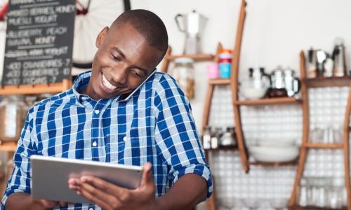 Café small business owner smiling and speaking on phone while using digital tablet