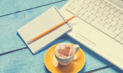 coffee with a heart in the milk on a teal table next to a notebook and a laptop