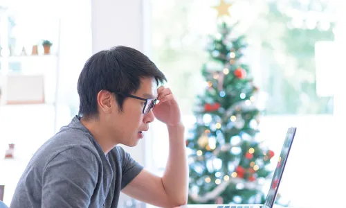 upset young main looks at his finances on laptop with Christmas tree in background