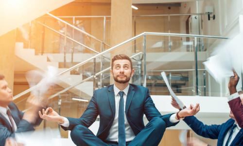 young businessman sits in meditation pose on table while colleagues shout and wave papers at him