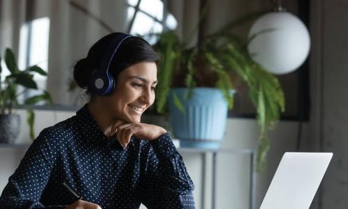 Young businesswoman takes notes while listening to headphones and watching laptop screen