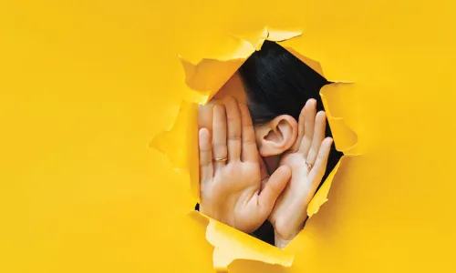woman holding cupping hands around ear to listen through hole in yellow paper background