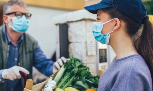 young woman in mask delivers box of groceries to elderly man during pandemic