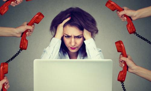 stressed businesswoman holding head while working on laptop surrounded by hands holding out many red telephone receivers