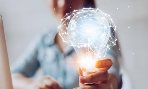 woman at desk holding out a glowing lightbulb with a network of data points forming the image of a brain on top of it