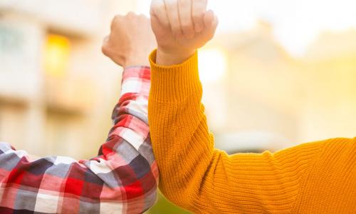 person in plaid shirt elbow bumps person in gold sweater in pandemic greeting