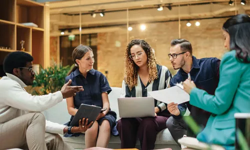 Group of individuals sitting around a coffee table looking at laptops and papers having a meeting